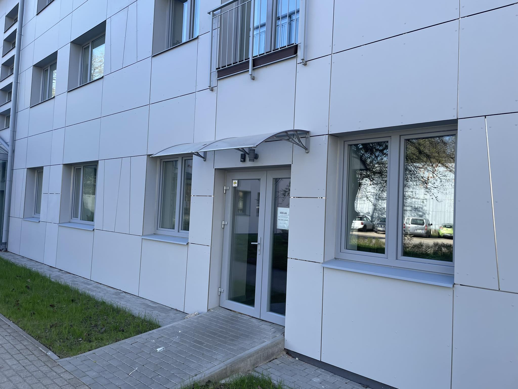 Attēlā redzama moderna ēkas fasāde ar gaišu apdari. Priekšplānā – stiklotas ieejas durvis ar nelielu jumtiņu virs tām un piekļuves celiņu. Blakus redzami logi, kuros atspoguļojas apkārtējā vide. Pie ēkas ir neliels zālājs un bruģēta taciņa.