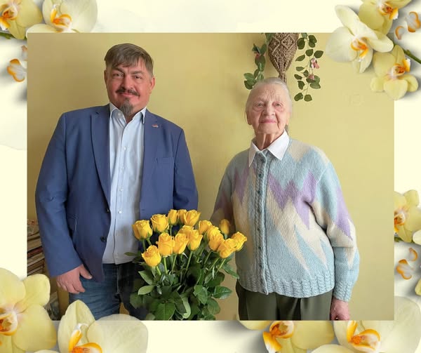 Attēlā redzami divi cilvēki – vīrietis un gados vecāka sieviete –, stāvot blakus telpā pie gaišas sienas. Vīrietis rokās tur lielu pušķi ar dzeltenām rozēm. Abi smaida. Sieviete ir sirmiem matiem, ģērbusies gaišā džemperī, vīrietis – zilā žaketē. Attēls dekorēts ar ziedu motīviem, radot svinīgu un sirsnīgu noskaņu.