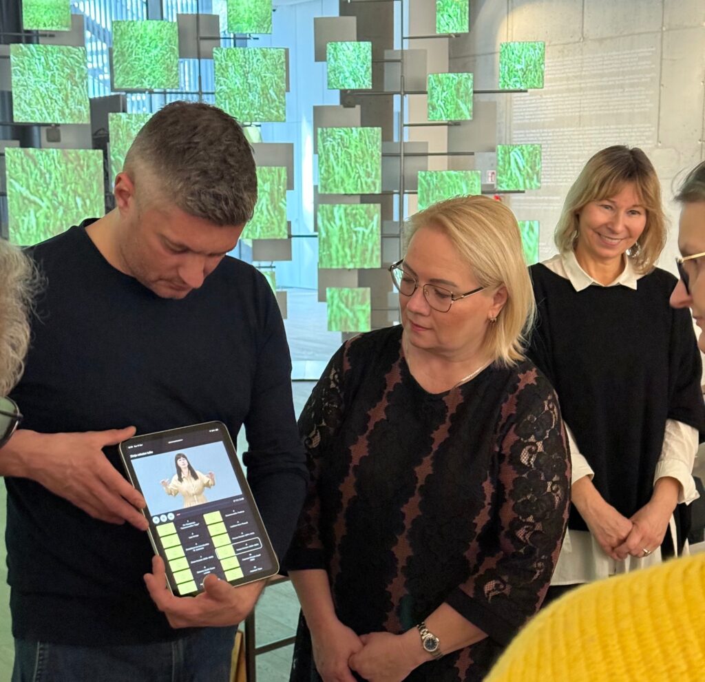 Attēlā redzami vairāki cilvēki stāvam modernā, gaišā telpā ar dekoratīviem, zaļiem kvadrātveida elementiem fonā. Priekšplānā vīrietis tur planšetdatoru un rāda tajā redzamu video ar sievieti, kas izmanto zīmju valodu. Blakus stāvoša sieviete uzmanīgi skatās uz ekrānu. Pārējie dalībnieki fonā vēro demonstrāciju. Atmosfēra ir ieinteresēta un izglītojoša, notiek digitāla risinājuma prezentācija.