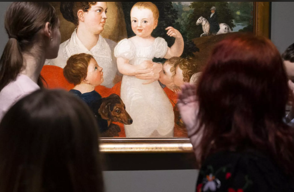 Cilvēki muzejā aplūko gleznu ar sievieti un bērniem. Gleznā redzama sieviete, kas tur mazuli rokās, apkārt ir vēl divi bērni un suns. Priekšplānā redzami apmeklētāji, kas stāv ar muguru pret skatītāju un vēro mākslas darbu.