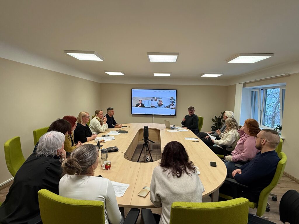 Plaša, gaiša sapulču telpa ar ovālu galdu, ap kuru sēž Latvijas Nedzirdīgo savienības domes locekļi domes sēdē. Ap galdu izvietoti aptuveni 13 cilvēki, daļa skatās uz runātāju, daļa – uz ekrānu telpas priekšā. Uz sienas redzams liels ekrāns ar attālināti pieslēgtiem dalībniekiem videozvanā. Uz galda – dokumenti, piezīmes, telefoni un kafijas krūzes. Telpa ir mierīga, darba atmosfēra, notiek diskusija.