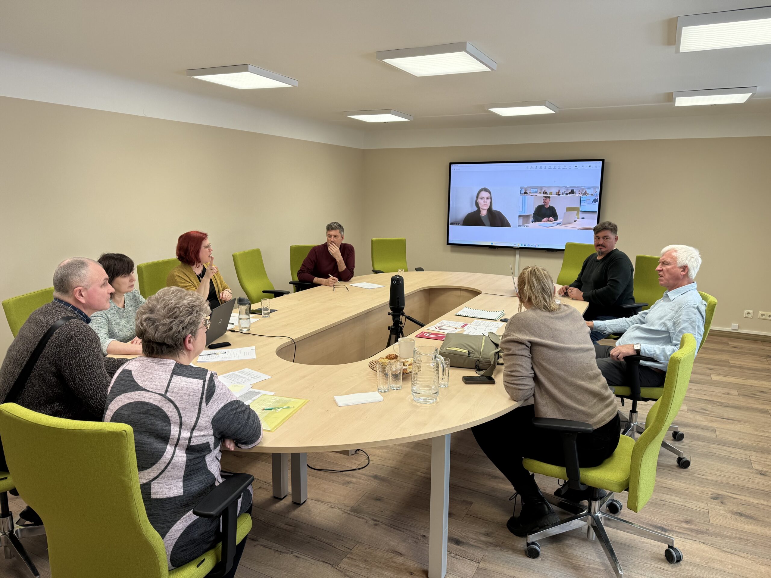 LNS valdes sēde sapulču telpā – ap ovālu galdu sēž valdes locekļi un kapitālsabiedrību pārstāvji, uz galda redzami dokumenti, piezīmes un ūdens glāzes. Daļa dalībnieku iesaistās diskusijā, citi klausās. Uz sienas ekrāna redzami attālināti pieslēgti dalībnieki videozvanā. Telpā valda mierīga, darba atmosfēra.