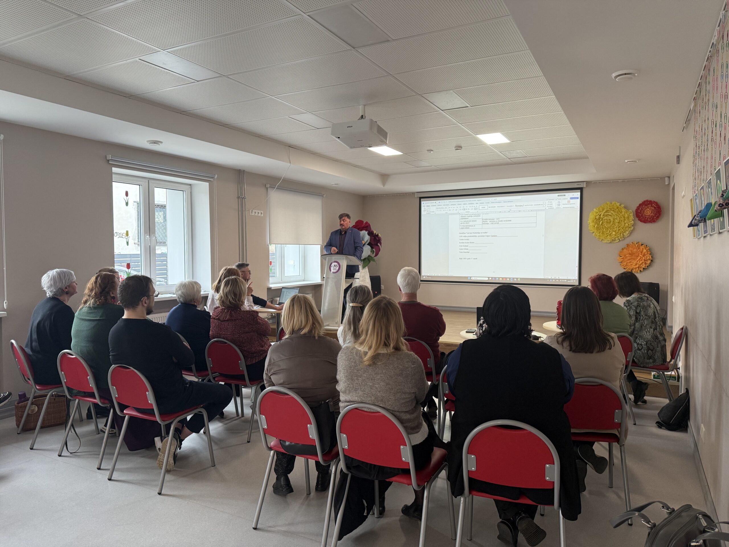 Attēlā redzama sapulce LNS telpās – cilvēki sēž rindās uz krēsliem un klausās runātāju, kurš stāv pie tribīnes telpas priekšā. Aiz runātāja uz ekrāna projicēts dokuments. Telpa ir gaiša, ar logiem kreisajā pusē, un dalībnieki vērsti uz priekšu, uzmanīgi sekojot prezentācijai.