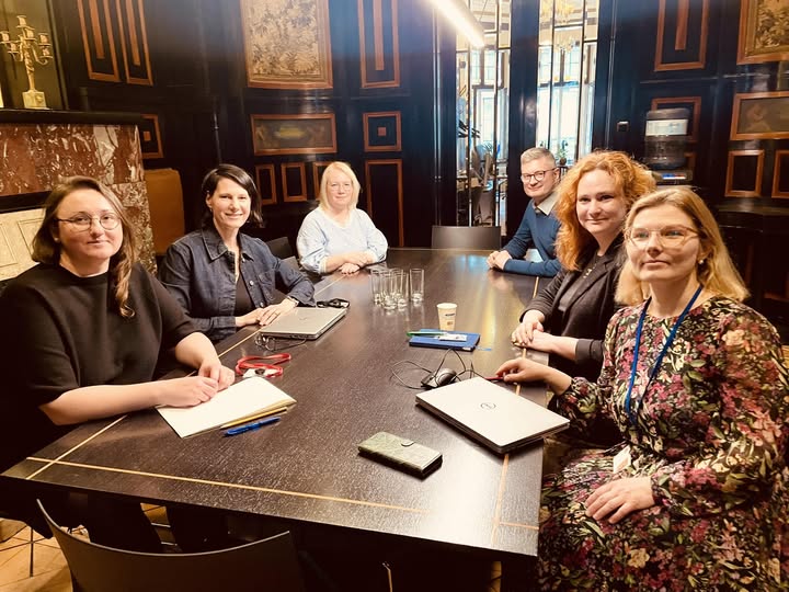 Attēlā redzami seši cilvēki, sēžot pie galda sapulču telpā ar tumšu, klasisku interjeru. Dalībnieki sēž ap galdu, uz kura novietoti datori, piezīmju bloki, telefoni un ūdens glāzes. Visi skatās kameras virzienā un smaida. Telpā redzami dekoratīvi sienu paneļi un gleznas, radot formālu un profesionālu tikšanās vidi.