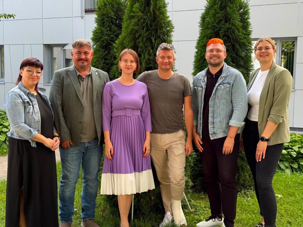 Grupas fotogrāfijā brīvā dabā pie ēkas redzami seši cilvēki, kuri stāv blakus un smaida kamerai.
