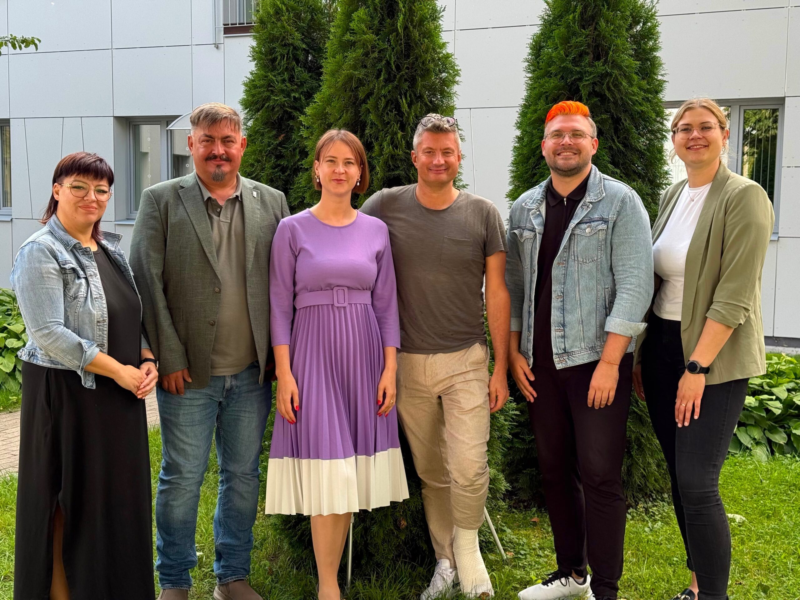 Grupas fotogrāfijā brīvā dabā pie ēkas redzami seši cilvēki, kuri stāv blakus un smaida kamerai.